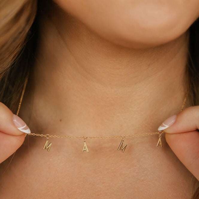 a woman is holding a gold mama dangle letter necklace made in canada