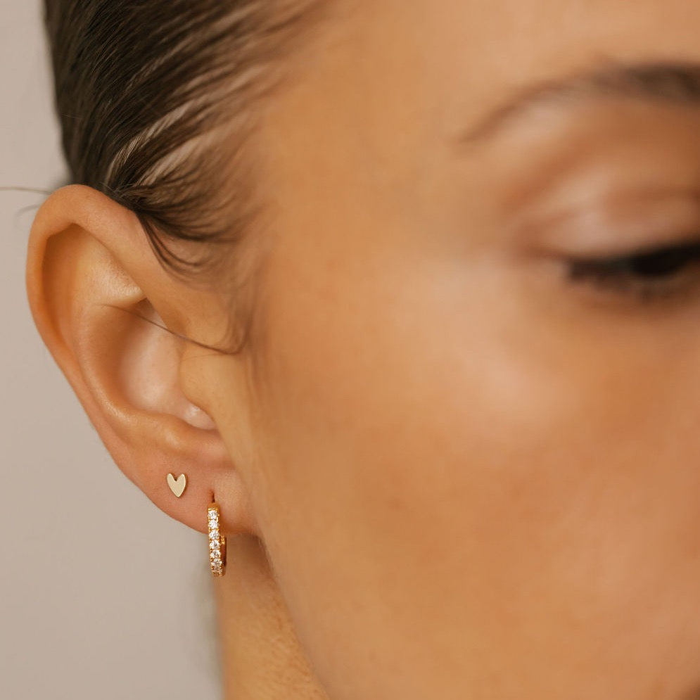 a woman is wearing gold heart stud earrings and pave diamond hoops made in Canada